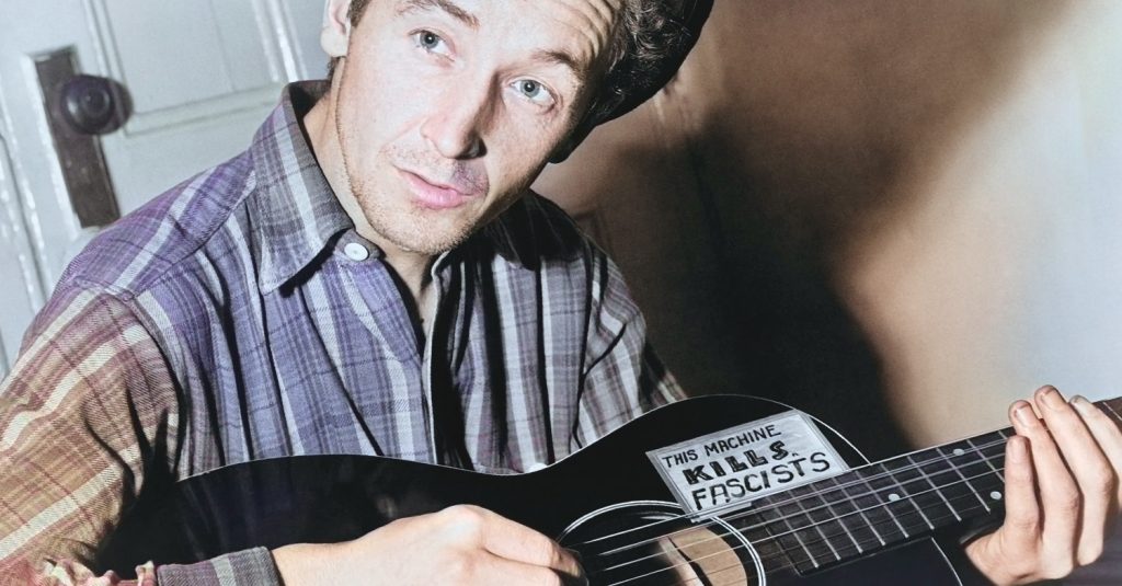 Woody Guthrie holding his guitar.  A sign on the guitar says, "This machine kills fascists." - colorized - Woody Guthrie Center